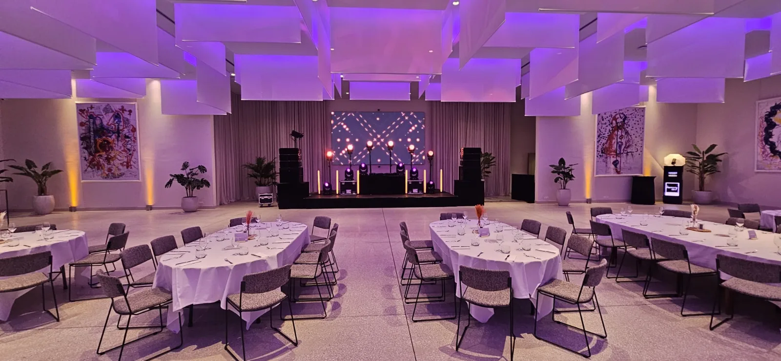Feestzaal in paars en warm licht met gedekte tafels en een DJ-podium met moving heads